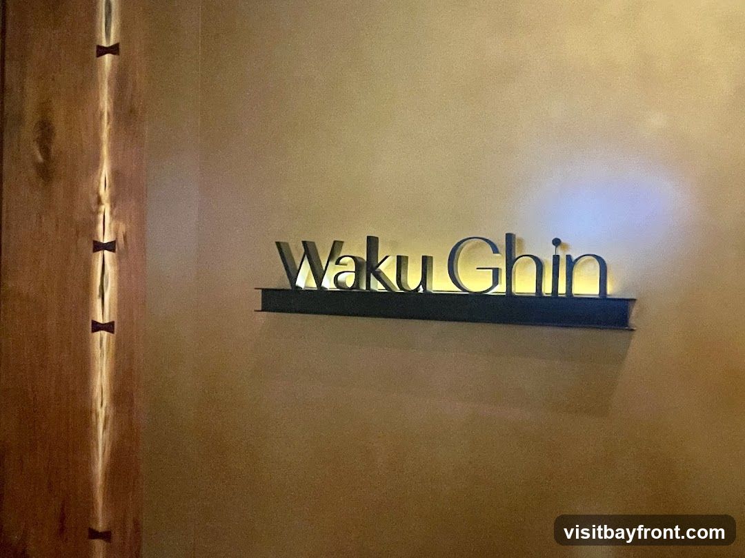 A close-up shot of the "Waku Ghin" sign mounted on a textured wall. The sign is made of dark, metallic letters with a warm, internal glow. To the left, a wooden panel with decorative inlays is visible.