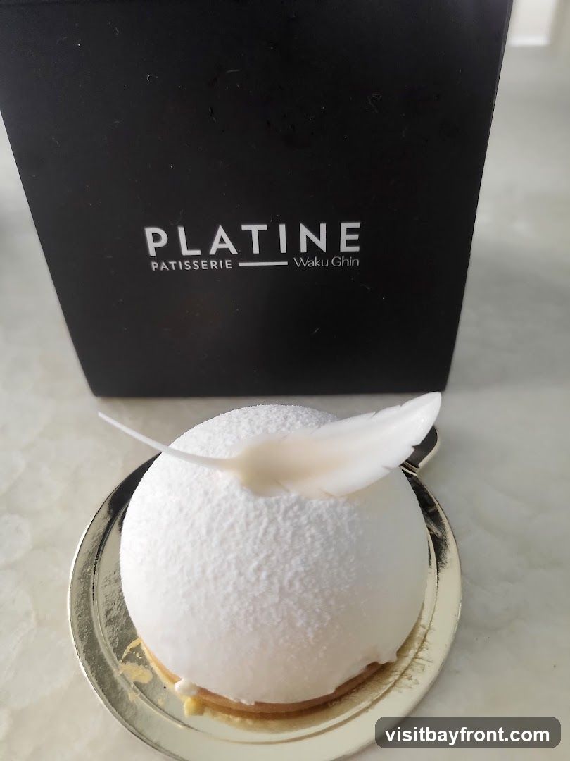 A white, spherical mousse cake sits on a gold foil-lined base. The cake is dusted with a fine white powder and adorned with a delicate white feather. Behind the cake, a black box with the logo "PLATINE PATISSERIE Waku Ghin" is visible.
