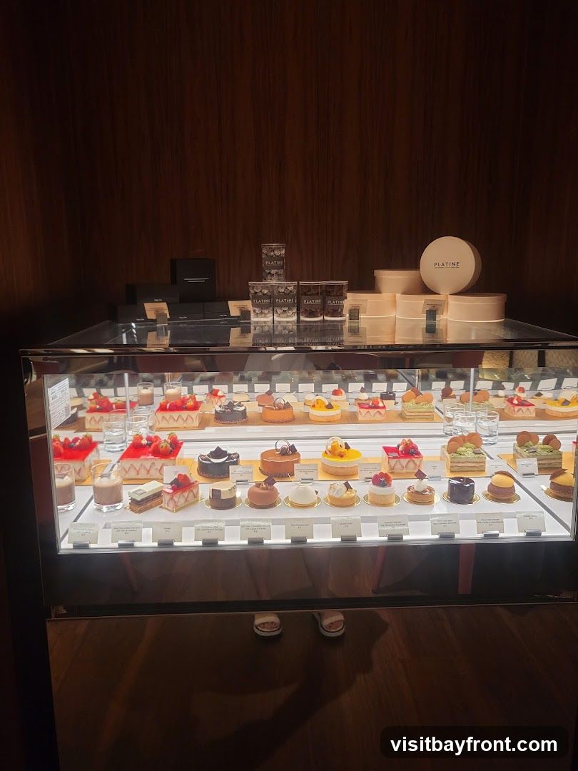 A refrigerated display case filled with a variety of small, artfully decorated cakes and pastries. The desserts are arranged on multiple tiers, with labels in front of each item. Above the display, there are boxes and round containers, some with the brand name "PLATINE" visible. The background is a dark, wood-paneled wall.