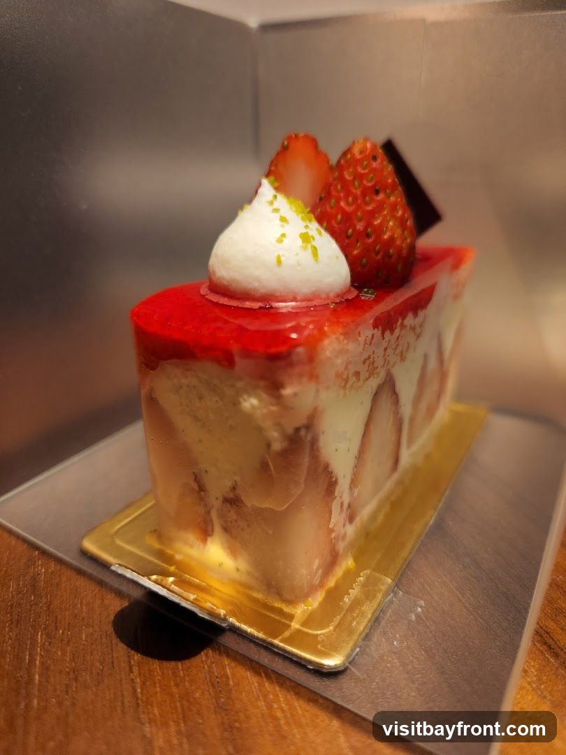 A slice of strawberry cake is presented on a golden cake board. The cake features layers of cream and strawberries, topped with a glossy red glaze, a dollop of whipped cream, fresh strawberries, and a piece of dark chocolate. The background is a metallic silver surface.