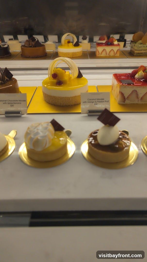 A display case filled with various pastries and cakes. In the foreground, two small tarts are visible, one topped with meringue and the other with a glossy caramel glaze and a white sphere. Behind them, larger cakes are arranged, including a strawberry shortcake and a yellow mousse cake with white chocolate decorations. Labels indicate some of the dessert names and prices.