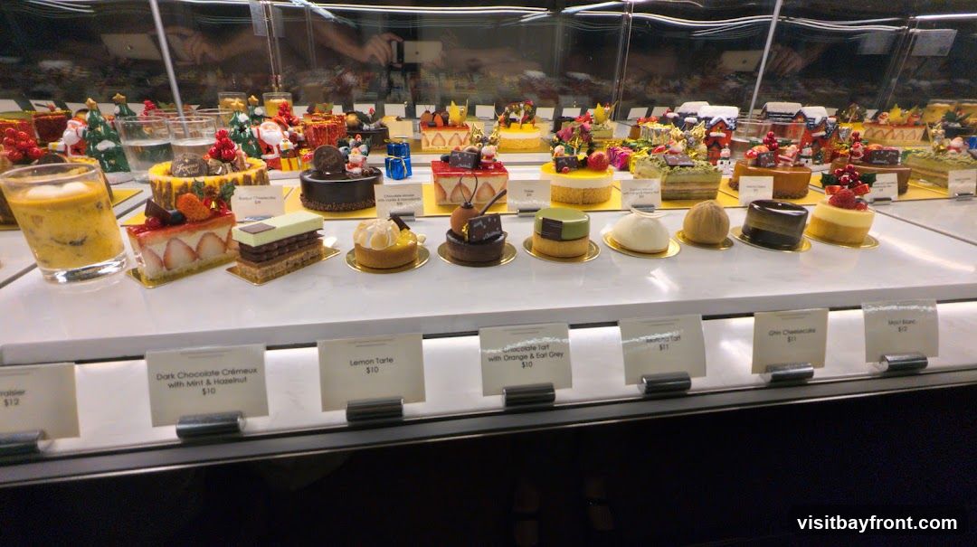 A display case filled with various pastries and cakes, many decorated with festive Christmas themes. The desserts include tarts, cheesecakes, and layered cakes. Labels with names and prices are visible in front of some items.