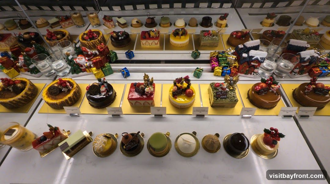 A display case filled with various Christmas-themed cakes and desserts. The top shelf shows smaller pastries and some drinks in glasses, decorated with Santa figures and gift boxes. The middle shelf features several individual cakes, including chocolate, strawberry, mango, and matcha flavors, also adorned with festive decorations. The bottom shelf presents a selection of smaller desserts and tarts.