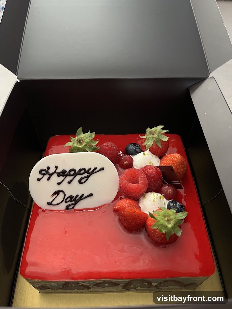 A square cake with a glossy red glaze is decorated with fresh strawberries, raspberries, blueberries, and grapes. A white oval plaque on the cake reads "Happy Day" in black script. The cake is presented in an open black box.