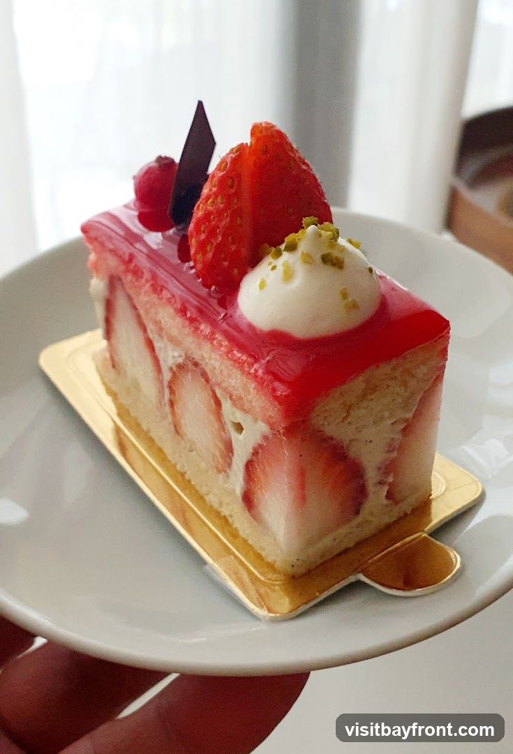 A slice of strawberry cake is presented on a white plate. The cake has layers of sponge cake and cream, filled with fresh strawberry slices. It is topped with a shiny red glaze, a dollop of whipped cream, chopped pistachios, a fresh strawberry half, a red berry, and a piece of dark chocolate.