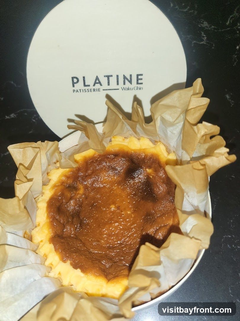 A close-up shot of a burnt Basque cheesecake, presented in a white container lined with brown parchment paper. The top of the cheesecake has a characteristic caramelized, slightly burnt appearance. A round, white lid with the embossed text "PLATINE PATISSERIE Waku Ghin" is visible in the background.