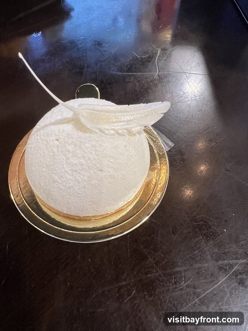 A close-up shot of a white dome-shaped cake with delicate white frosting and a decorative white feather-like garnish. The cake sits on a gold cake board, which is placed on a dark, reflective surface.