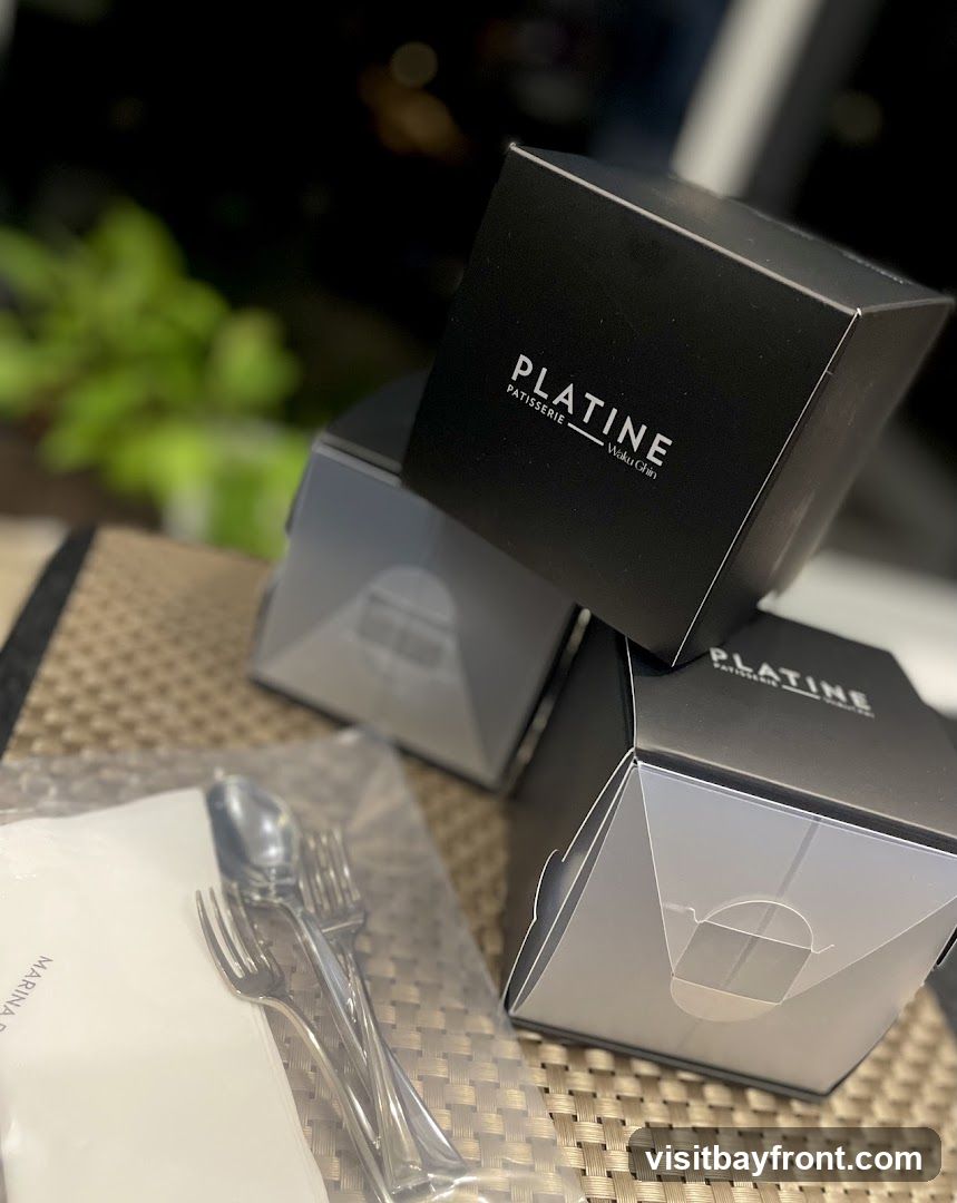 Three black takeaway boxes with the logo "PLATINE PATISSERIE Waku Ghi" are stacked on a table. In the foreground, a set of cutlery (fork and spoon) is wrapped in plastic next to a white napkin with the text "MARINA BAY" visible. The background is blurred, showing some greenery and lights.