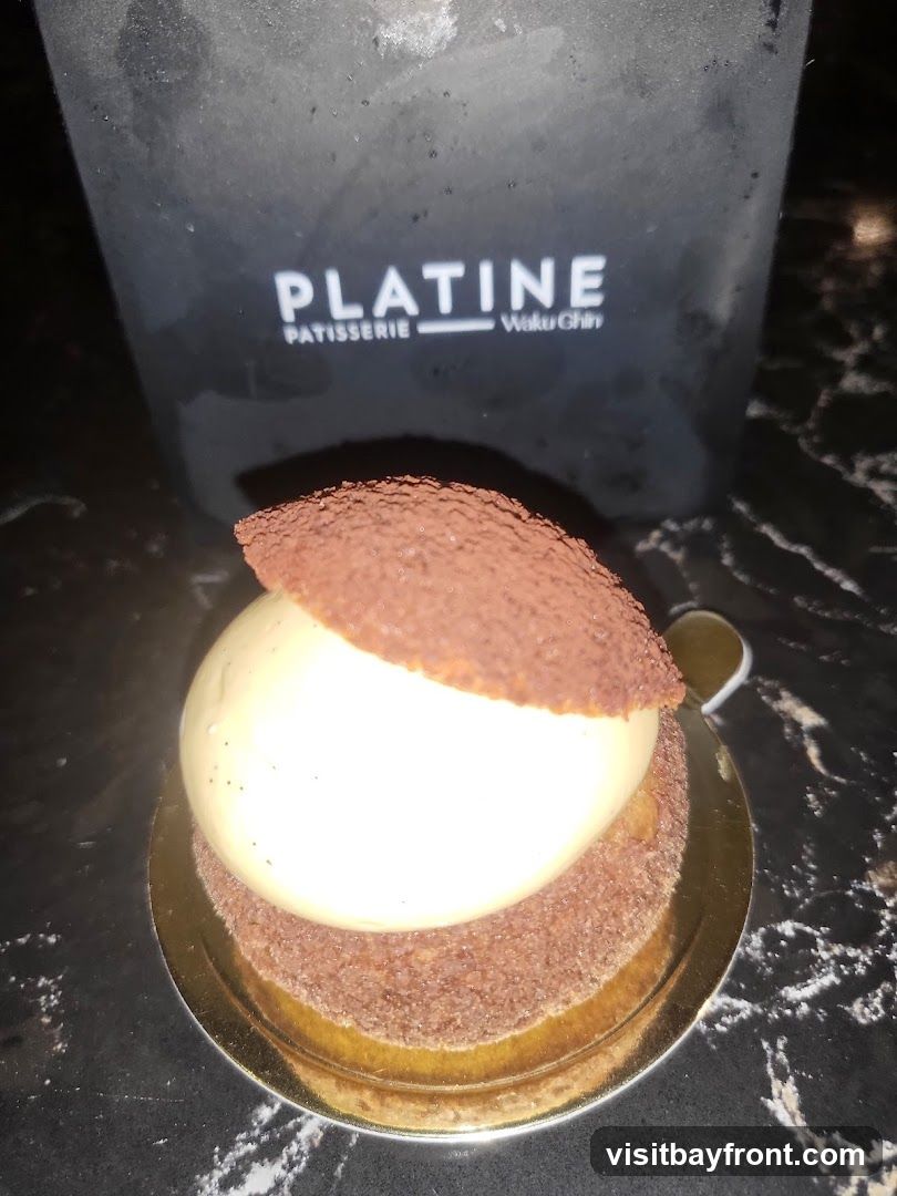 A close-up of a dessert, possibly a mousse or cake, with a white creamy top and a chocolate-dusted base. Behind it, a black container or bag with the white text "PLATINE PATISSERIE Waku Chin" is visible. The dessert is on a gold circular base, placed on a dark, marbled surface.