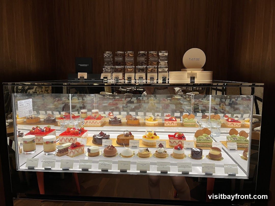 A refrigerated display case filled with various pastries and cakes. The pastries are arranged on multiple tiers and include items topped with fruits like strawberries, as well as chocolate and cream-based desserts. Some items are presented in individual portions, while others are larger cakes. Labels with names and prices are visible in front of some items. Above the display case, there are boxes of cookies and a round box with the logo "PLATINE". The background is a dark wood-paneled wall.
