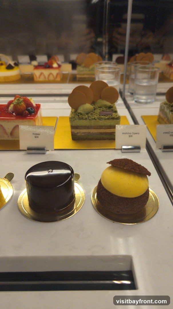 A display case filled with various pastries and cakes. In the foreground, there are two round pastries, one dark chocolate and one with a yellow dome. Behind them, a rectangular cake with green layers and a strawberry-topped cake are visible. Labels indicate "Fraisier $30" and "Matcha Opera $30". In the background, more desserts and glasses of water are displayed.