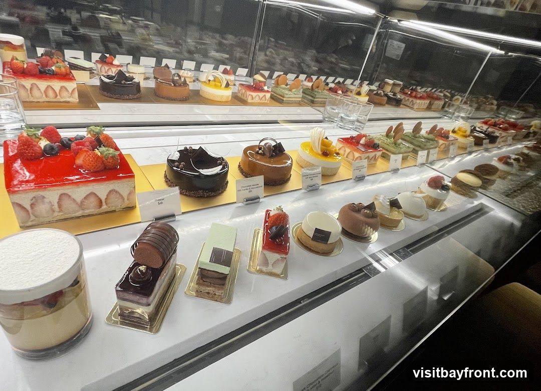 A refrigerated display case filled with a variety of beautifully decorated cakes and pastries. The desserts include fruit-topped cakes, chocolate mousse cakes, and layered pastries. Some items have small labels with prices.