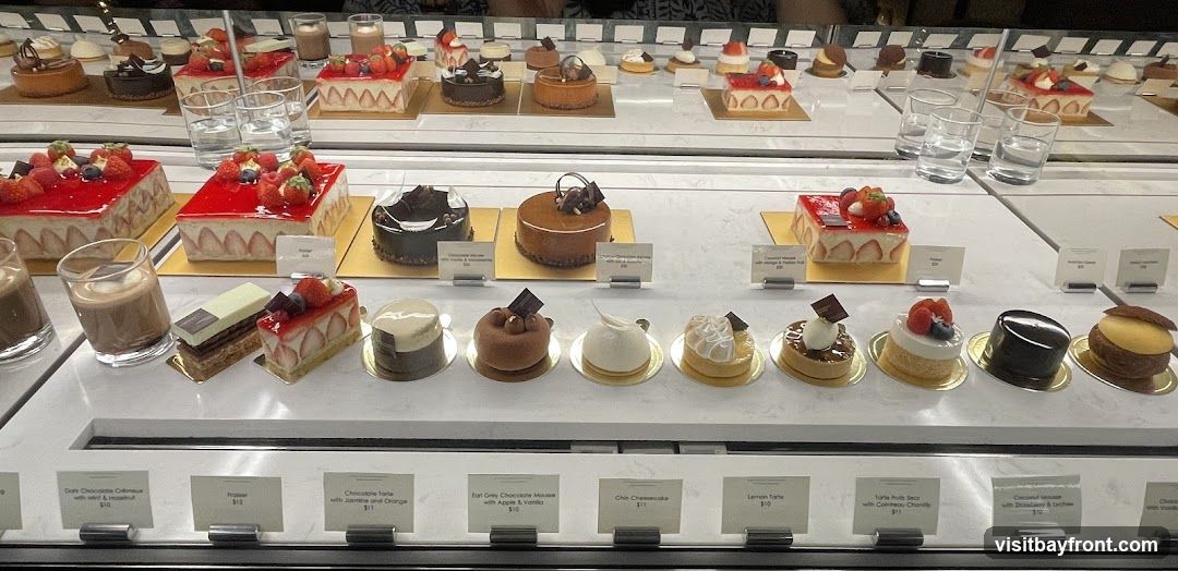 A display case filled with various beautifully crafted desserts, including cakes, tarts, and mousse cups. Many of the desserts are topped with fresh fruits like strawberries and raspberries. Small descriptive cards are placed in front of some of the items.