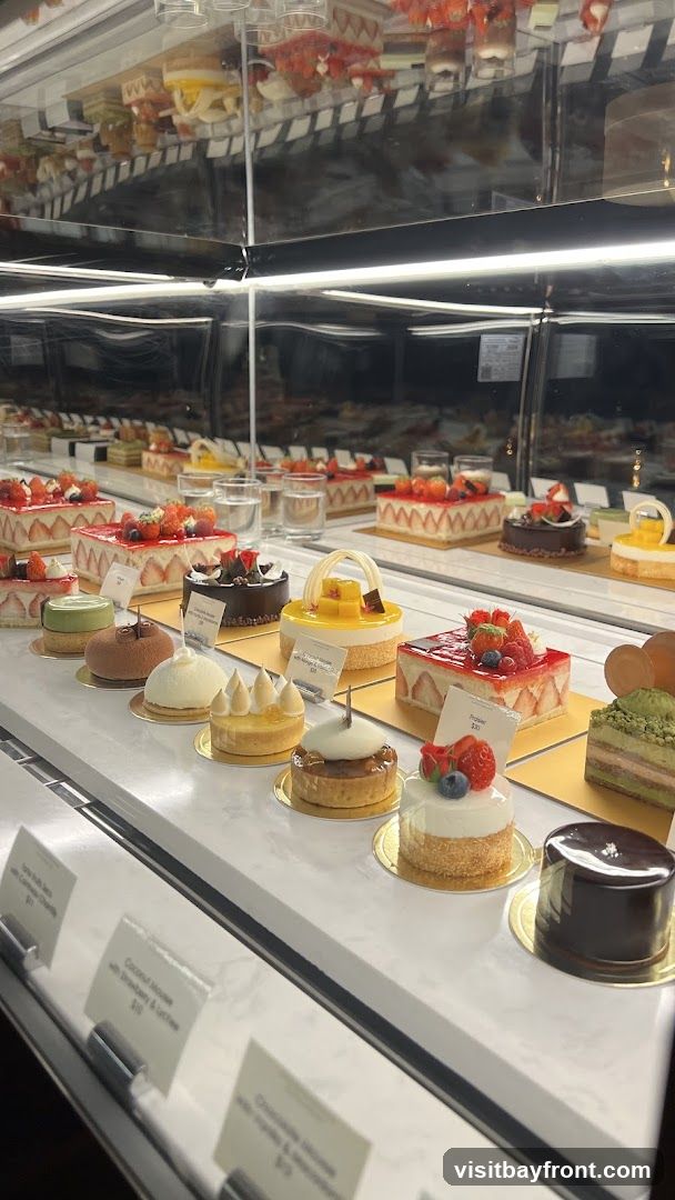A display case filled with various beautifully decorated cakes and pastries. The desserts include fruit tarts, mousse cakes, and layered cakes, many topped with fresh berries and fruits. Small labels with descriptions and prices are visible in front of some items.