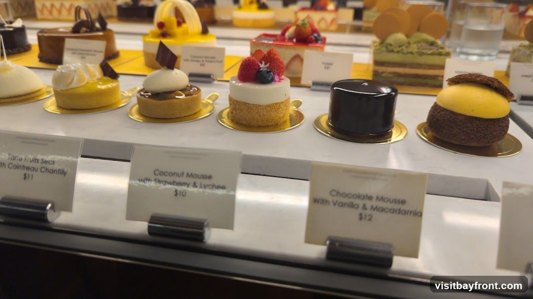 A display case filled with various small cakes and pastries. In the foreground, there are three small signs with descriptions and prices of some of the desserts. The desserts include a tart with fruit, a white mousse cake with berries, and a dark chocolate mousse cake. In the background, more elaborate cakes are visible.
