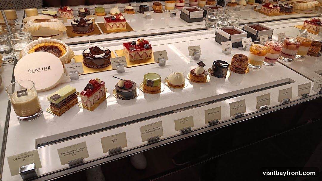 A display case filled with various beautifully crafted pastries and desserts. In the foreground, there are layered cakes, tarts, and individual desserts in glasses. A round box with the logo 'PLATINE PATISSERIE' is also visible. The desserts include items like strawberry tarts, chocolate cakes, and layered parfaits. Small labels with names and prices are placed in front of some of the desserts.