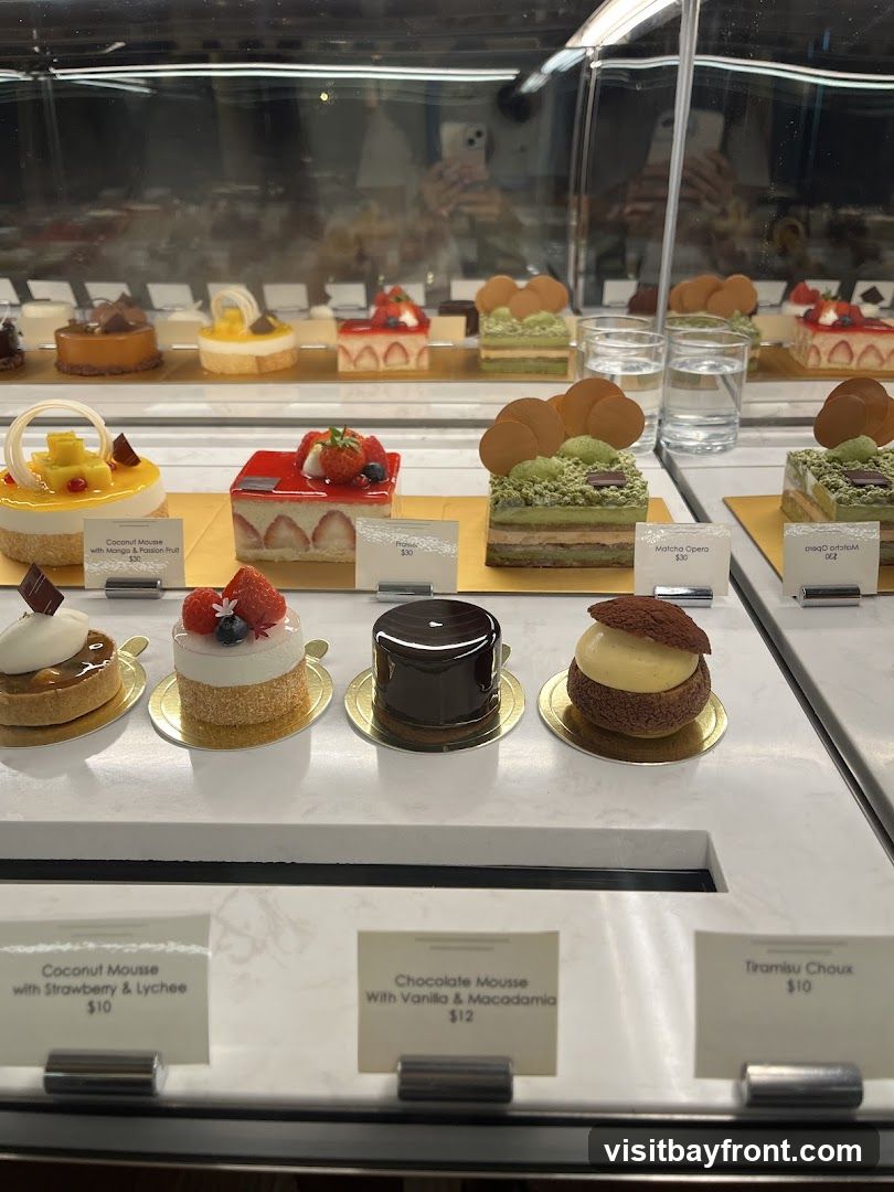 A display case filled with various pastries and cakes. In the foreground, there are several individual desserts with labels. Visible labels include 'Coconut Mousse with Strawberry & Lychee $10', 'Chocolate Mousse With Vanilla & Macadamia $12', and 'Tiramisu Choux $10'. In the background, more desserts are visible, including layered cakes and tarts.