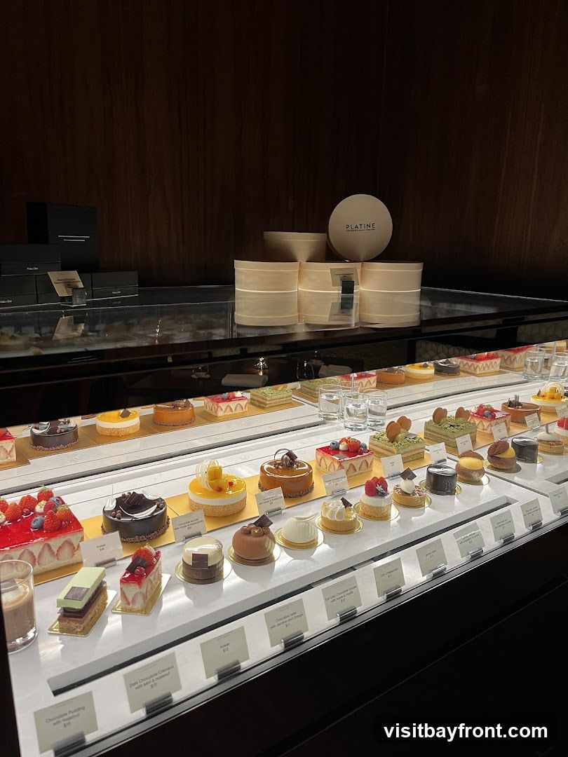 A display case filled with various beautifully decorated pastries and cakes. The pastries are arranged on multiple tiers, showcasing a variety of flavors and designs, including fruit-topped cakes, chocolate creations, and tarts. Some pastries have small labels with their names and prices. In the background, there are boxes and round containers, one with the logo 'PLATINE'.