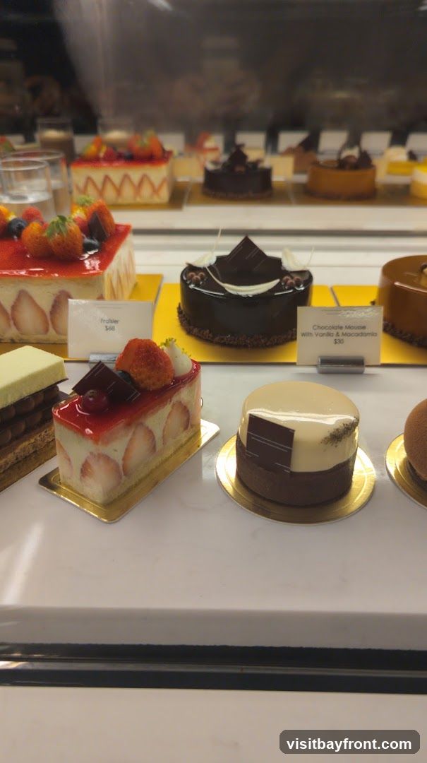 A display case filled with various pastries, including fruit tarts, chocolate cakes, and mousse desserts. Some pastries are topped with fresh strawberries and blueberries. Labels indicate some of the dessert names and prices.