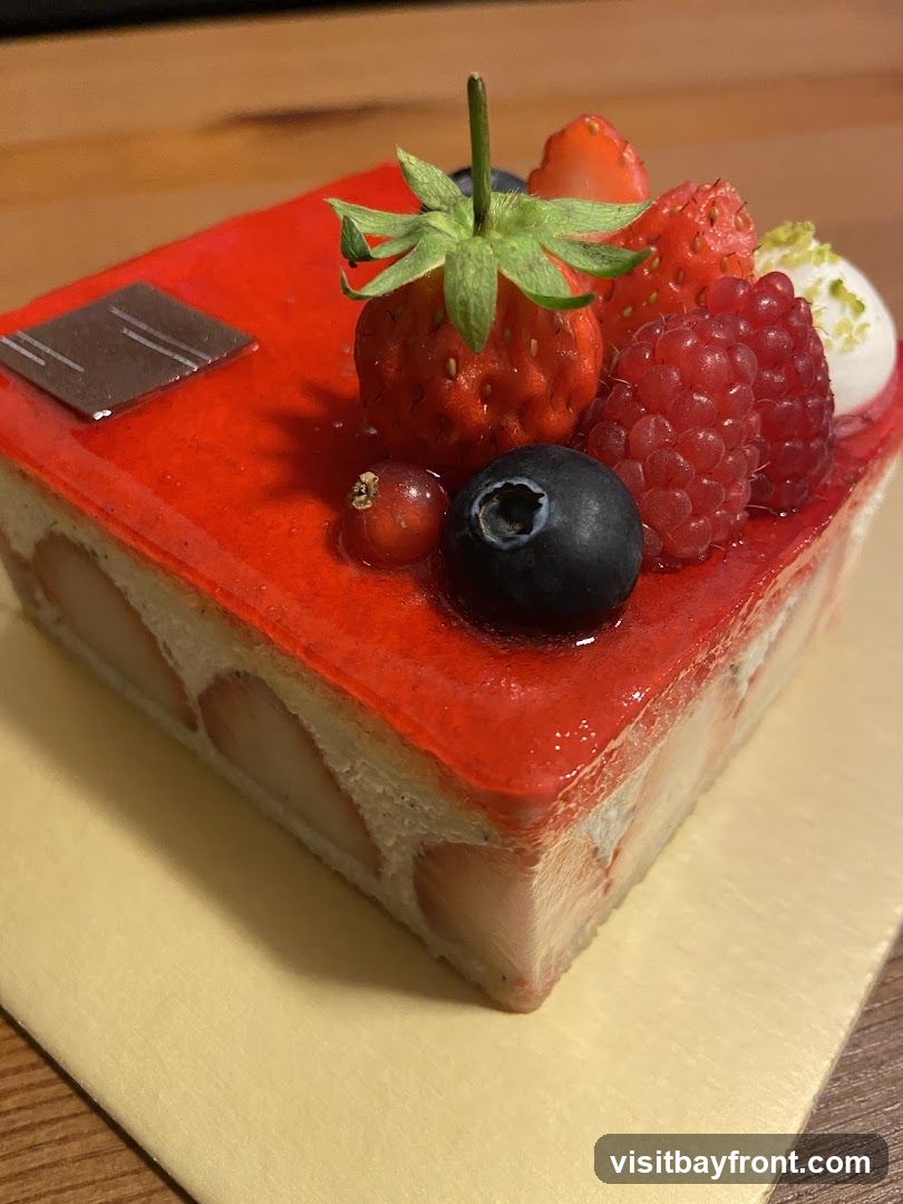 A square cake with a bright red glaze, topped with fresh strawberries, raspberries, blueberries, and a red currant. The cake layers are visible on the side, showing sliced strawberries embedded in cream. A small square of dark chocolate is placed on the top left corner.