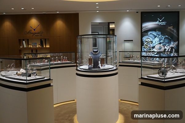 The interior of a luxury watch store, featuring multiple glass display cases showcasing watches and jewelry. A large screen displays intricate watch mechanisms. The store has a sophisticated and modern design with wooden paneling and soft lighting.