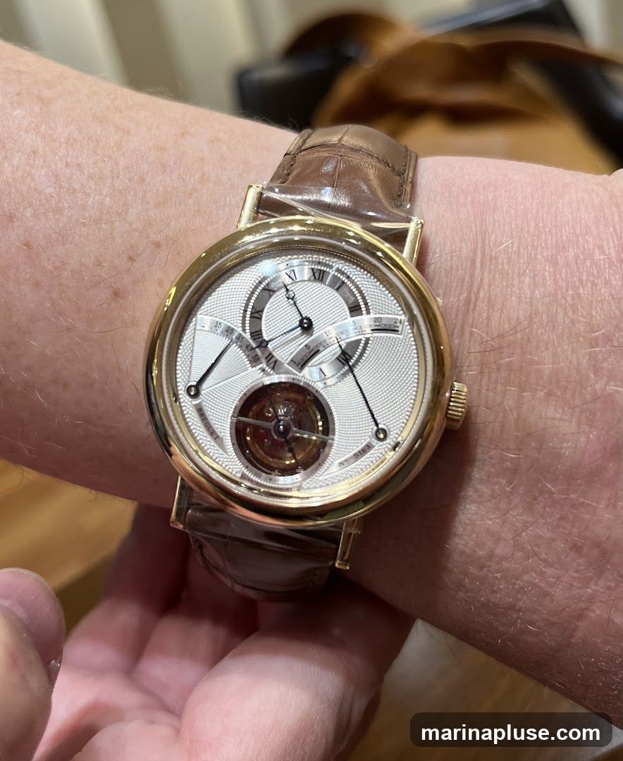 A close-up shot of a gold watch with a white dial and Roman numerals on a person's wrist. The watch has a brown leather strap. The dial features a tourbillon complication at the 6 o'clock position and sub-dials for time and power reserve.