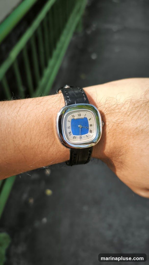 A close-up shot of a person's wrist wearing a watch with a silver-toned square case and a black leather strap. The watch face has Roman numerals and a blue rectangular accent in the center. The background is blurred, showing a green railing and a dark, textured surface.