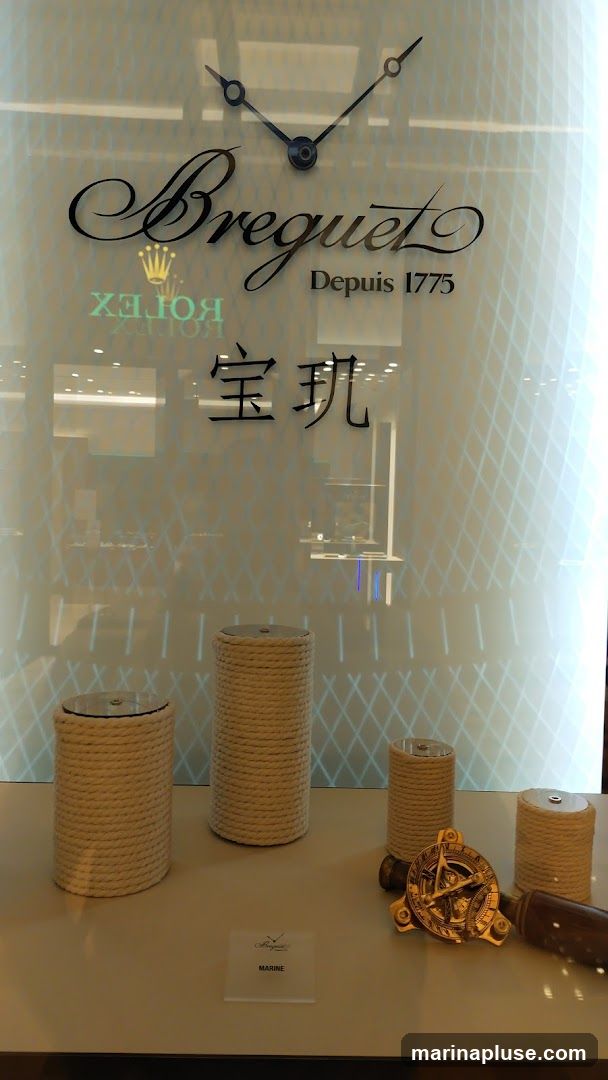 A display window featuring the Breguet logo and clock hands. Below the logo, the text "Depuis 1775" is visible, along with Chinese characters. The display includes several cylindrical stands wrapped in rope, a brass sundial, and a wooden handle. A small sign reads "MARINE" with the Breguet logo. Reflections of a Rolex logo are also visible on the glass.