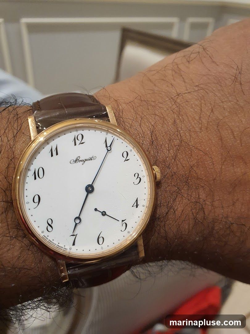 A close-up shot of a man's wrist wearing a gold Breguet watch with a white dial and a brown leather strap. The watch face displays Arabic numerals and has a second sub-dial. The watch is partially covered by protective plastic.