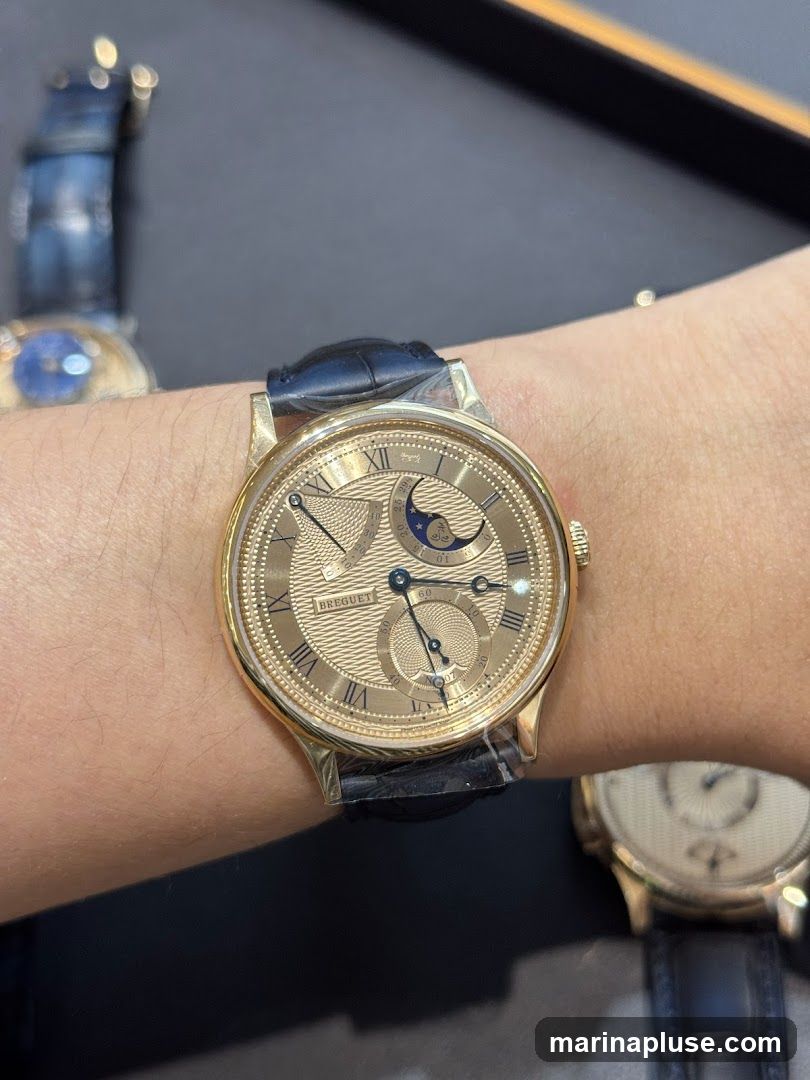 A close-up shot of a person's wrist wearing a gold watch with a black leather strap. The watch face is intricately detailed with Roman numerals, a guilloché pattern, and a moon phase complication. Another watch is visible in the blurred background.