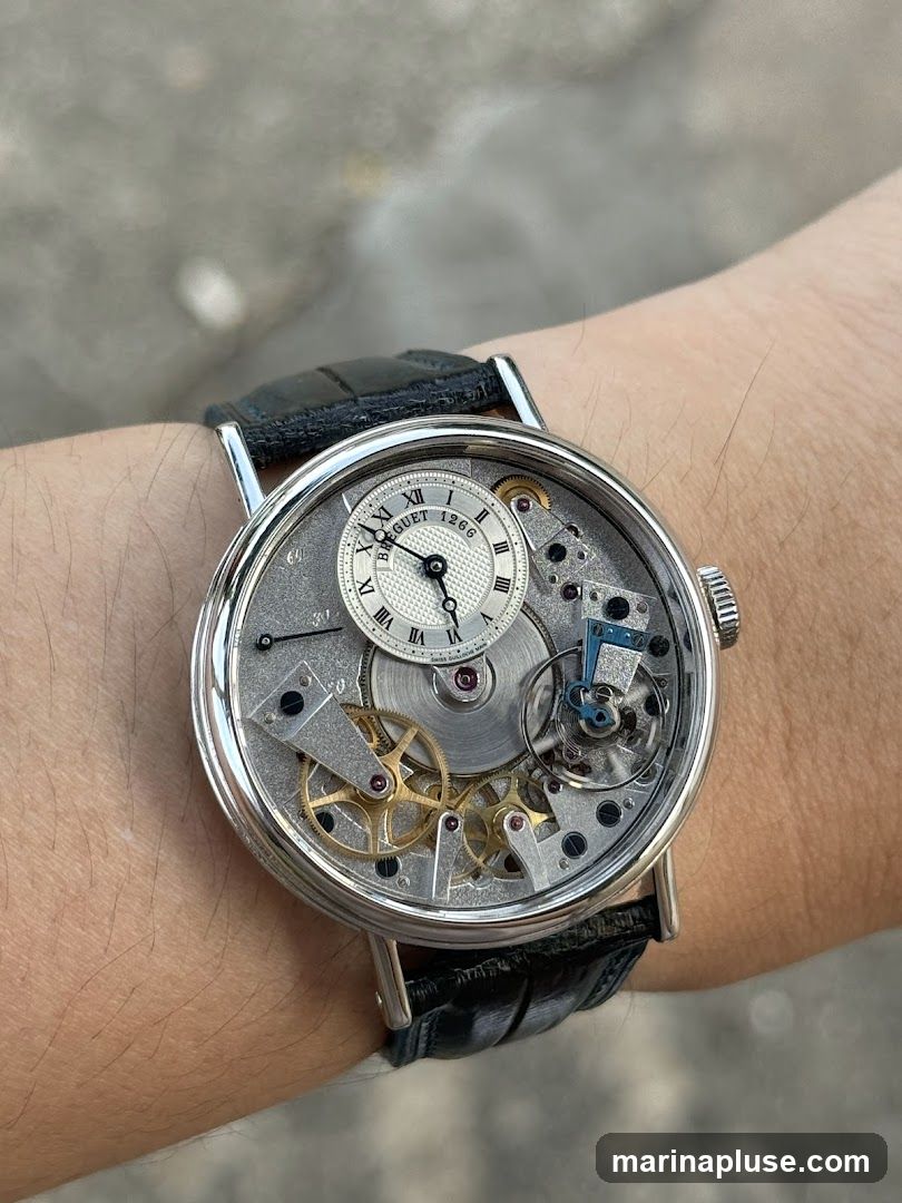 A close-up shot of a Breguet watch on a person's wrist. The watch features an intricate mechanical movement with visible gears and components. The dial is white with Roman numerals and a sub-dial for seconds. The watch has a black leather strap.