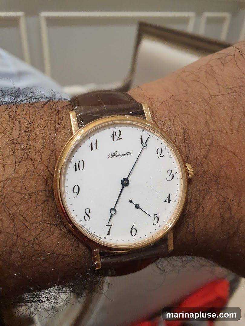 A close-up shot of a gold Breguet watch with a white dial and Roman numerals on a person's wrist. The watch has a brown leather strap. The background is blurred, showing hints of furniture and possibly a room interior.