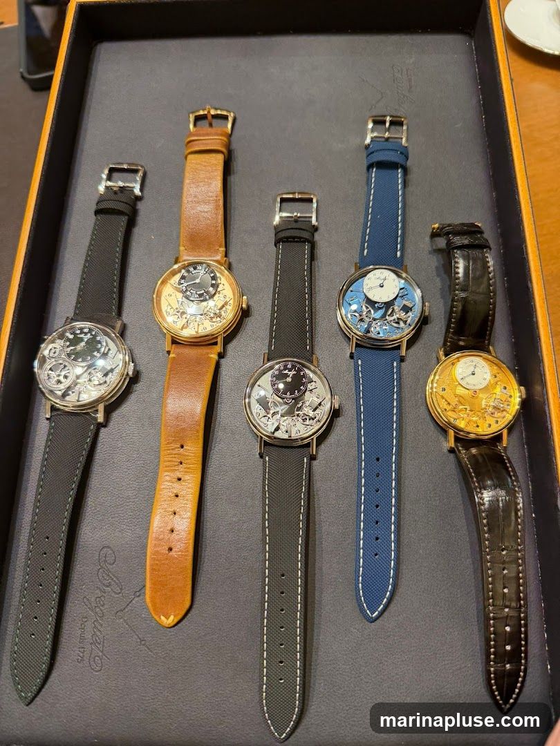A close-up overhead view of six luxury watches laid out in a dark tray. The watches have intricate, exposed mechanical movements and come with various strap colors including black, tan, blue, and dark brown alligator leather. The tray has a subtle embossed logo that appears to be "Breguet".