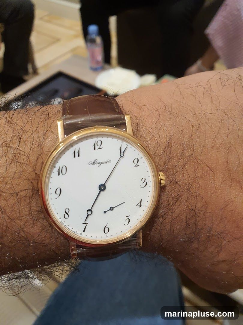 A close-up shot of a person's wrist wearing a gold Breguet watch with a brown leather strap. The watch face is white with black Roman numerals and hands. In the background, out of focus, are people sitting around a table with a bottle of water and some food.