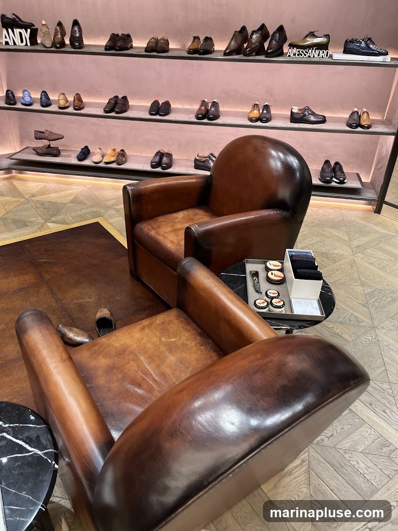 A well-lit shoe store interior featuring multiple shelves displaying various men's dress shoes. The shelves are organized by style and color, with some shoes presented on individual stands. In the foreground, two distressed leather armchairs are arranged on a patterned rug, suggesting a comfortable waiting or browsing area. A small round table next to one of the chairs holds shoe care products and a small stack of papers. The store's branding, "ANDY" and "ALESSANDRO," is visible on the shelving units.