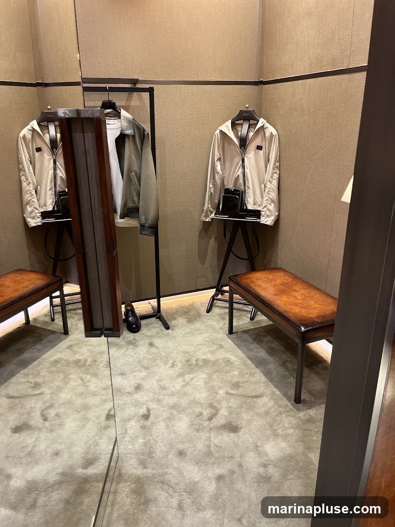 A fitting room with two jackets on display, each on a stand with a small bag. There are two leather benches and a mirror. A single black shoe is visible on the floor.