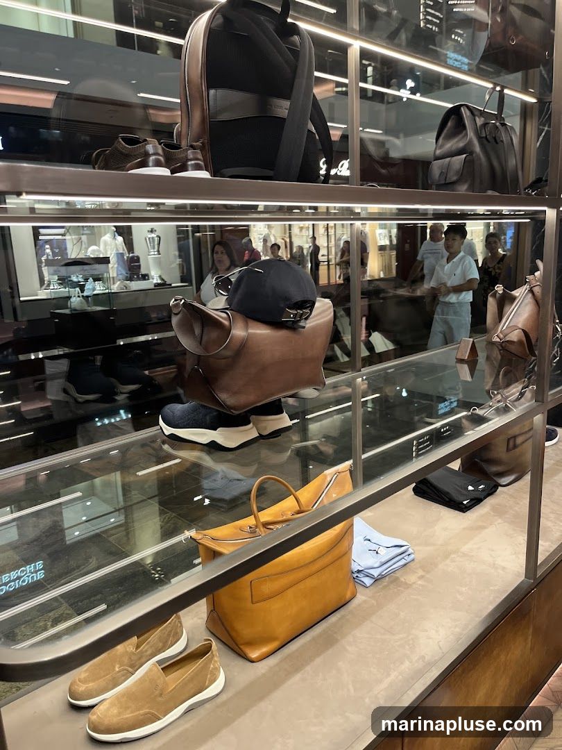A display case filled with various leather goods and accessories. On the top shelf, a black backpack and a pair of brown shoes are visible. The middle shelf features a brown leather saddle bag with a black cap on top, and a pair of blue sneakers. The bottom shelf showcases a large tan leather duffel bag, a pair of tan loafers, and folded light blue shirts. In the background, people are browsing the store, and reflections of other displays can be seen.
