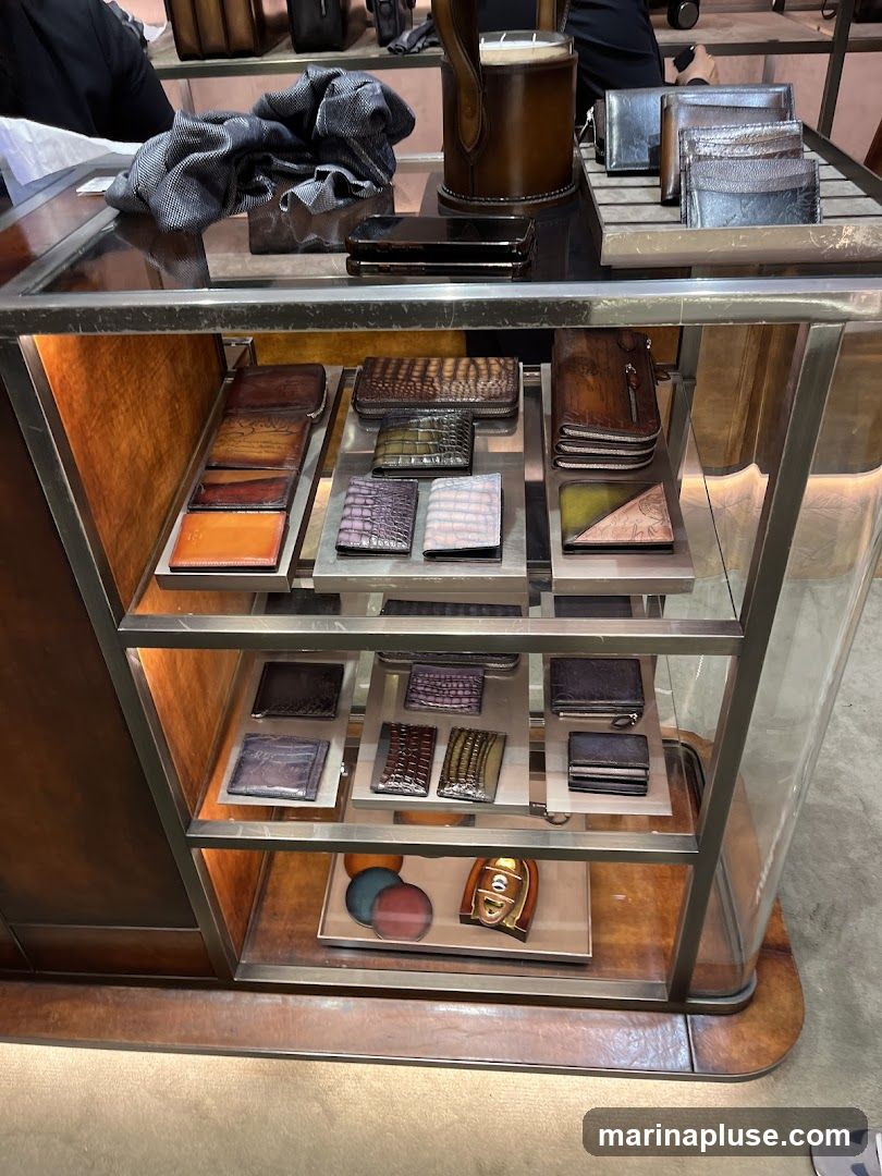 A display case filled with various leather wallets and accessories. The items are arranged on shelves within a glass-enclosed cabinet. The products include wallets in different colors and textures, such as crocodile and patterned leather. Some items are folded, while others are open. The display also features a few round leather swatches and a decorative metal emblem. The lighting highlights the rich tones of the leather.