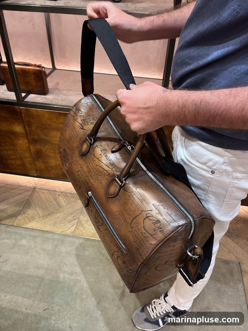 A man is holding a brown leather duffle bag with a script pattern. The bag has two top handles and a detachable shoulder strap. In the background, there are shelves displaying other leather goods. The man is wearing white pants and a dark blue patterned shirt. He is also wearing grey and navy sneakers.