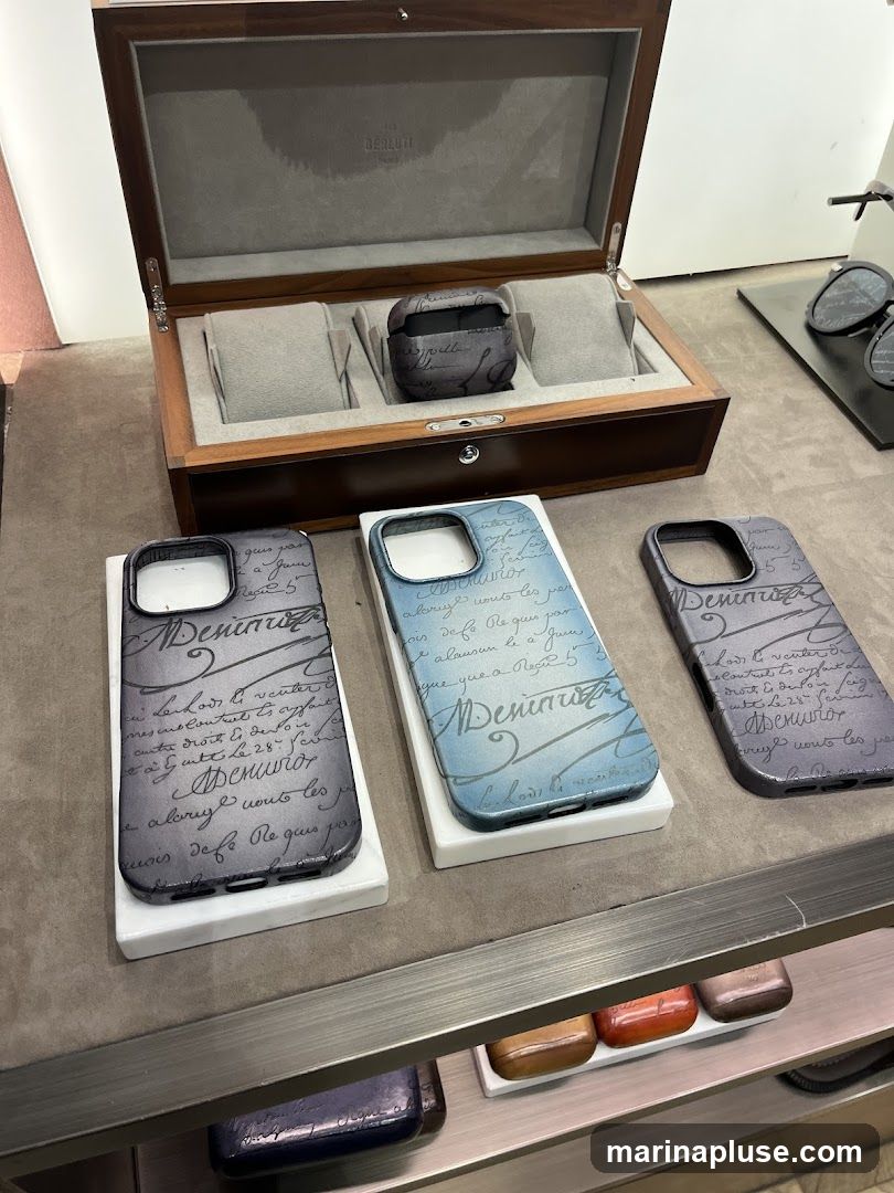 A display of Berluti accessories including phone cases, an AirPods case, and a watch box. The phone cases and AirPods case feature a script pattern. The watch box is made of wood and has the Berluti logo inside.