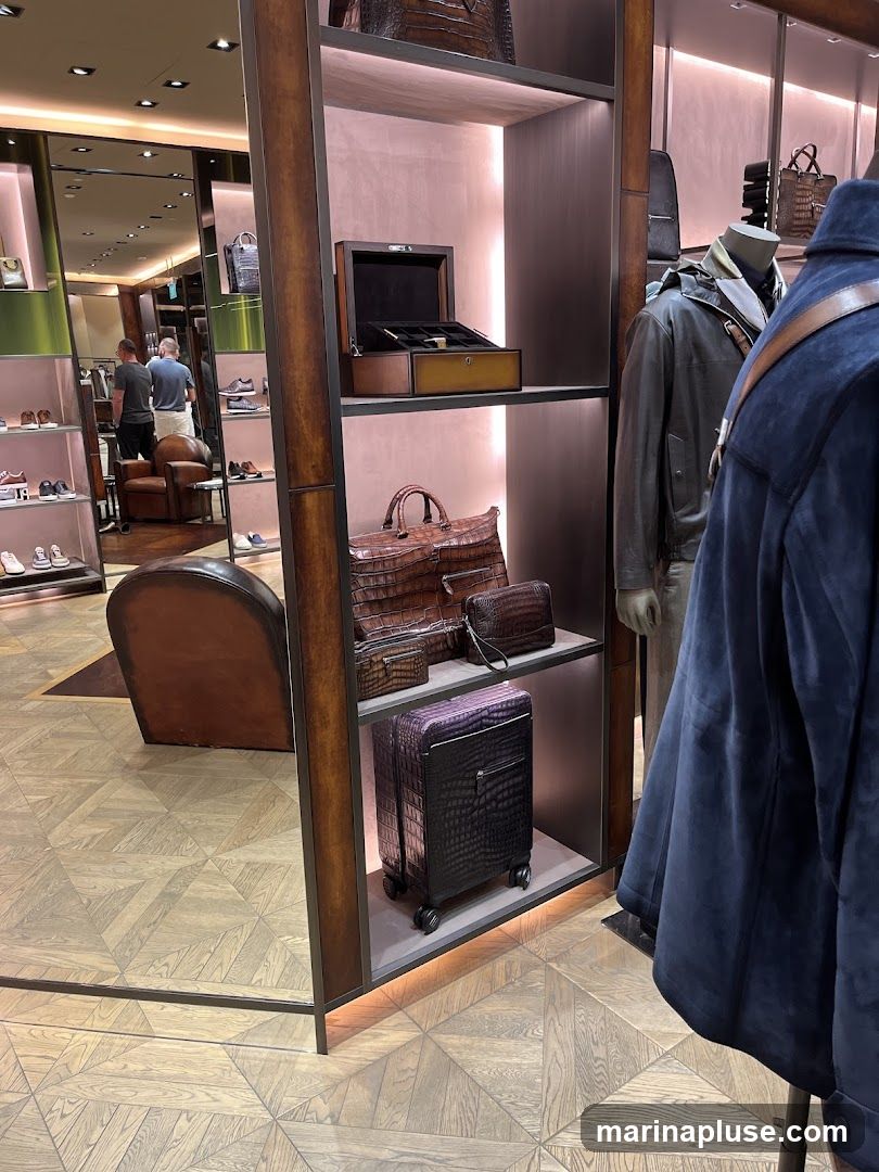 A retail display featuring luxury leather goods, including handbags, a jewelry box, and a suitcase, all with a crocodile-embossed texture. Mannequins dressed in outerwear are also visible. In the background, a reflection shows a store interior with shelves of shoes and two men browsing.