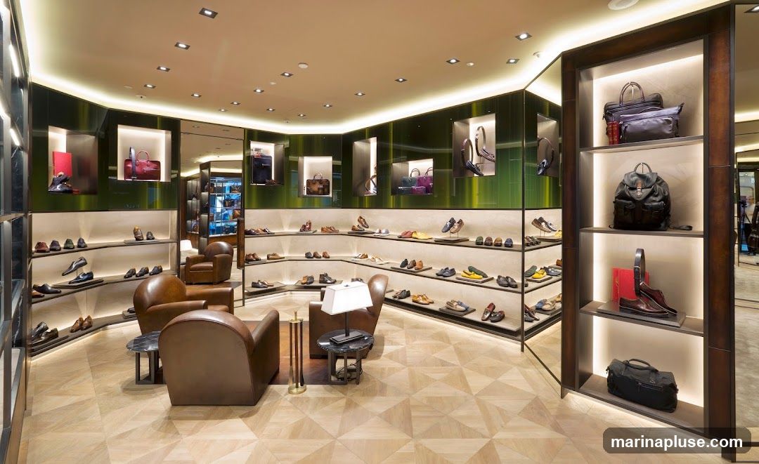 A well-lit, modern interior of a luxury store showcasing a wide selection of men's and women's footwear and handbags. The display shelves are arranged in a curved fashion, featuring shoes neatly placed on tiered shelves. Several handbags and backpacks are displayed in illuminated niches and on vertical shelving units. Comfortable leather armchairs are placed in the seating area, with a small coffee table between them. The flooring is a light-colored wood with a geometric pattern.