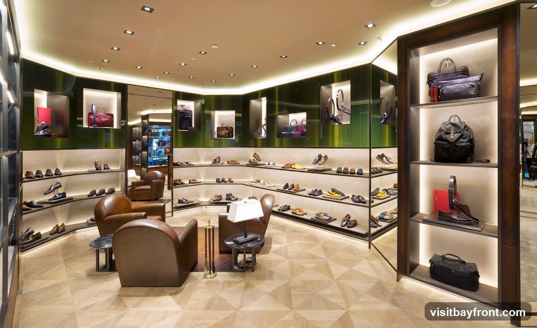 A well-lit, modern interior of a luxury store showcasing a wide selection of men's and women's footwear and handbags. The display shelves are arranged in a curved fashion, featuring shoes neatly placed on tiered shelves. Several handbags and backpacks are displayed in illuminated niches and on vertical shelving units. Comfortable leather armchairs are placed in the seating area, with a small coffee table between them. The flooring is a light-colored wood with a geometric pattern.