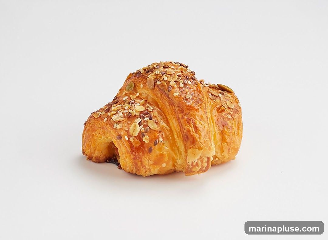 A single golden-brown croissant topped with a mix of seeds and oats, photographed against a plain white background.