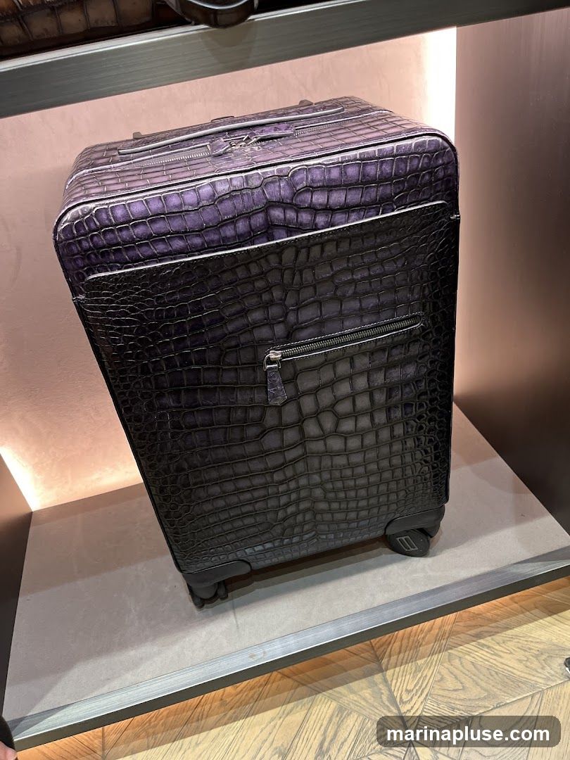 A close-up shot of a rolling suitcase with a crocodile-embossed pattern. The suitcase is primarily black, with a gradient effect transitioning to a deep purple on the top half. It features zippered compartments and sits on a display shelf with a wooden floor visible below.