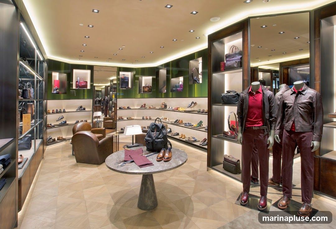 The interior of a high-end menswear store, featuring displays of shoes, handbags, and apparel on mannequins. The store has a modern and luxurious design with polished floors, ambient lighting, and mirrored walls. Leather armchairs are placed in a seating area.
