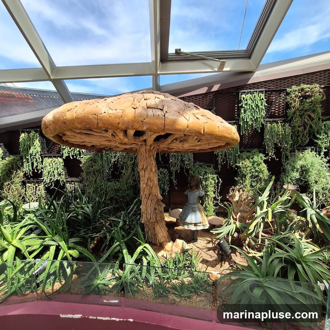 A large, rustic wooden mushroom sculpture is the centerpiece of a botanical display. Behind it, a small statue of Alice in Wonderland stands amongst lush greenery. The display is set against a wall covered in hanging plants and is illuminated by natural light filtering through a glass ceiling.