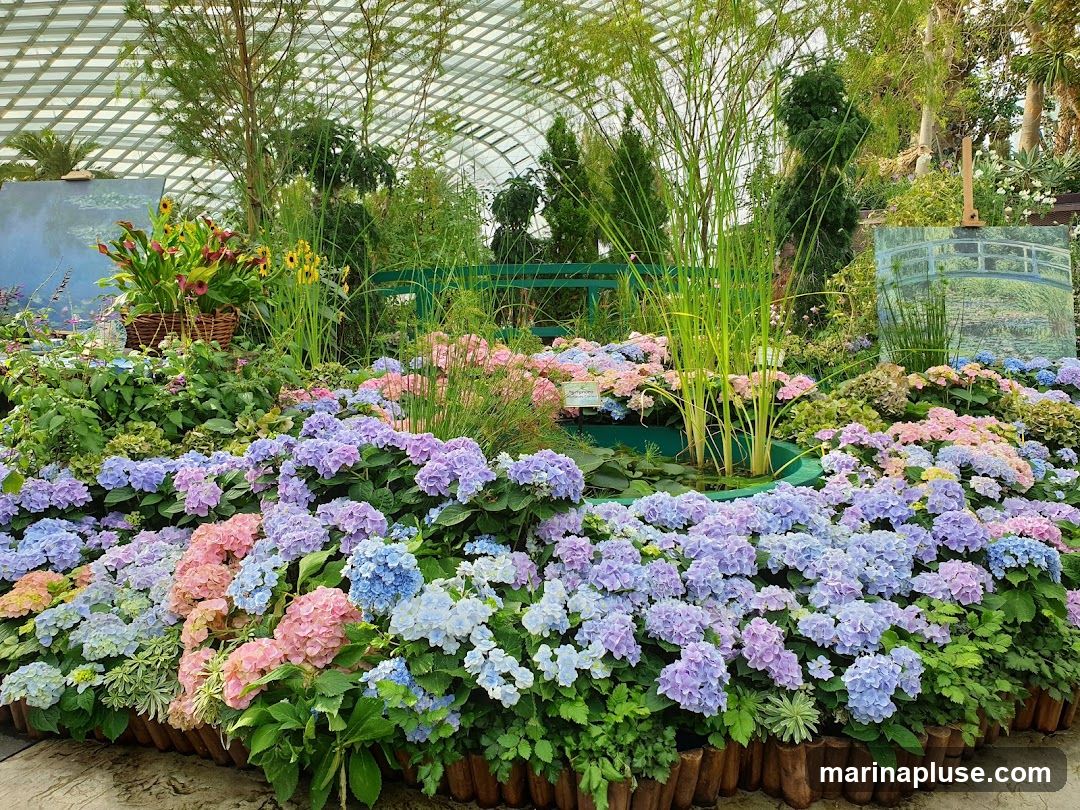 A vibrant display of hydrangeas in shades of pink, blue, and purple fills the foreground, interspersed with lush green foliage. Tall grasses rise from a central pond, adding vertical interest. In the background, two paintings are displayed on easels, one depicting water lilies and a bridge, reminiscent of Monet's work, and another with calla lilies and sunflowers in a woven basket. The scene is set within a greenhouse with a distinctive arched, grid-like ceiling.