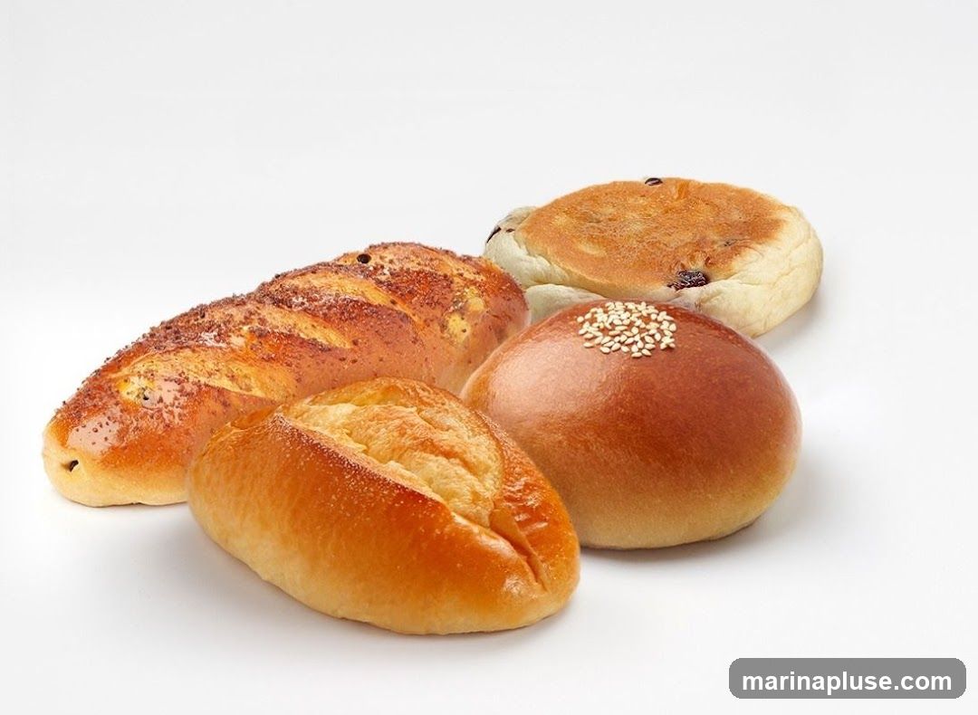 Four types of baked goods on a white background: a long loaf with dark speckles, a small oval bun with sesame seeds on top, a round golden-brown bun, and a flat circular bread with raisins. All appear freshly baked with shiny crusts.