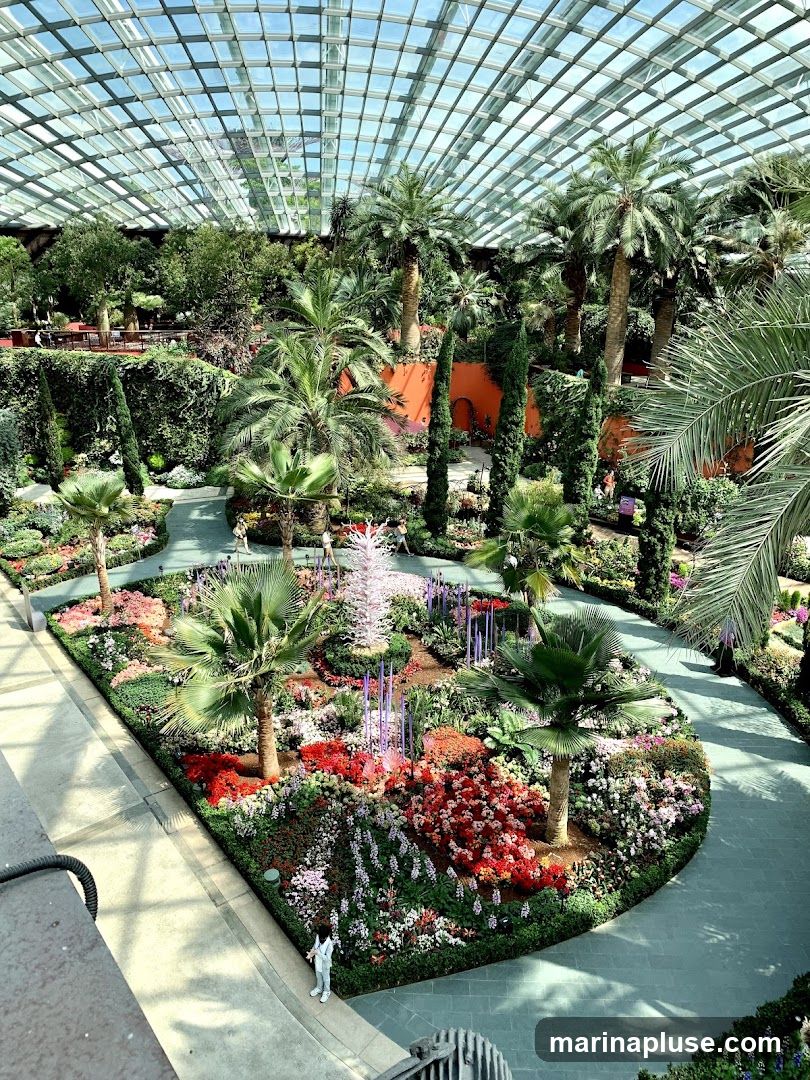 A vast indoor botanical garden with a glass ceiling, featuring a variety of palm trees, lush greenery, and a vibrant flower bed in the center. The flower bed is a colorful display of red, pink, and white flowers, with purple decorative elements and a tall, white, sculptural piece. Pathways wind through the garden, and a few visitors can be seen exploring the space.